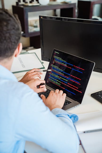 A person with short dark hair, wearing a light blue shirt, is seated at a desk working on a laptop. The laptop screen displays lines of colored programming code, featuring syntax highlighted with brig