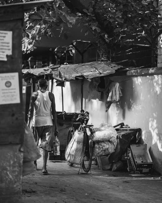 A person walking down a narrow alleyway with a bicycle parked along the side. The bicycle is loaded with large bags and bundles of waste materials, including plastic and fabric, which are secured to i