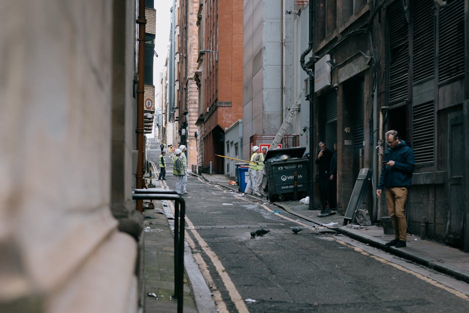 A person walking down a narrow alleyway with a bicycle parked along the side. The bicycle is loaded with large bags and bundles of waste materials, including plastic and fabric, which are secured to i