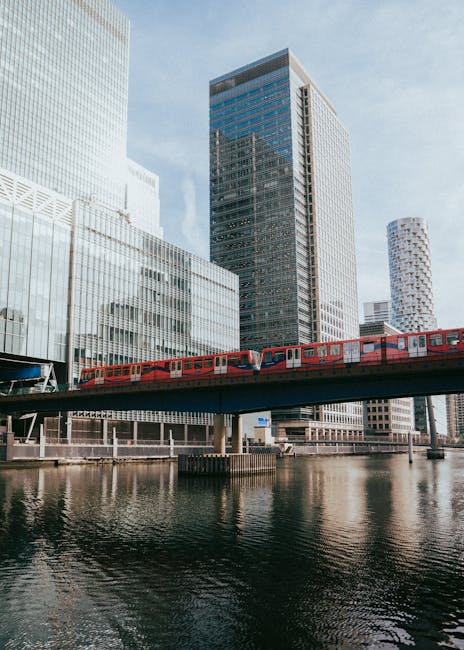 A modern red train is seen moving along an elevated railway track across a body of water in an urban setting. In the background, there are several high-rise buildings with glass facades reflecting the
