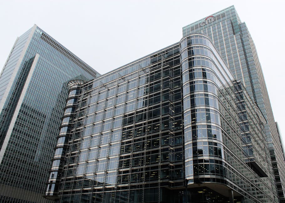 A modern red train is seen moving along an elevated railway track across a body of water in an urban setting. In the background, there are several high-rise buildings with glass facades reflecting the